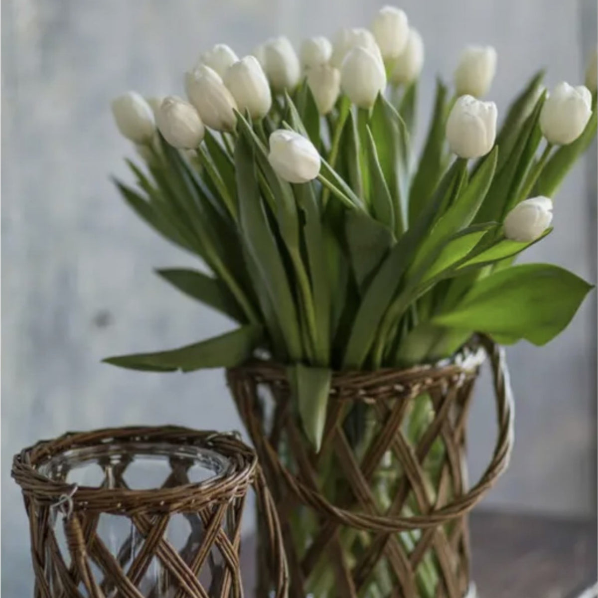 A rustic woven willow vase elegantly displayed with a bouquet of white tulips. This charming vase features a sturdy glass insert and a woven wicker frame, making it a perfect centerpiece for farmhouse or boho-style home décor. Measures 7.5 inches in diameter and 9 inches in height.