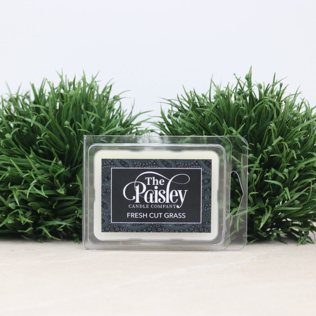 A 2.5 oz clamshell-style wax melt labeled “The Paisley Candle Company – Fresh Cut Grass,” displayed upright between two faux grass accents on a light-colored surface.