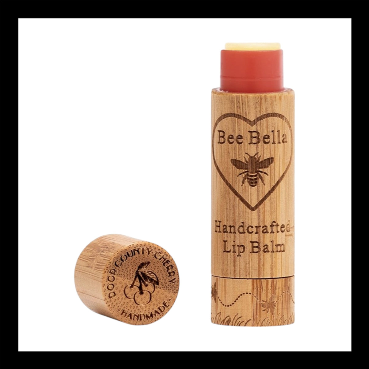 Bee Bella Handcrafted Lip Balm in eco-friendly bamboo packaging, featuring an engraved bee and heart design. The cap, placed beside the tube, is engraved with 'Door County Cherry Handmade' alongside a cherry illustration. 